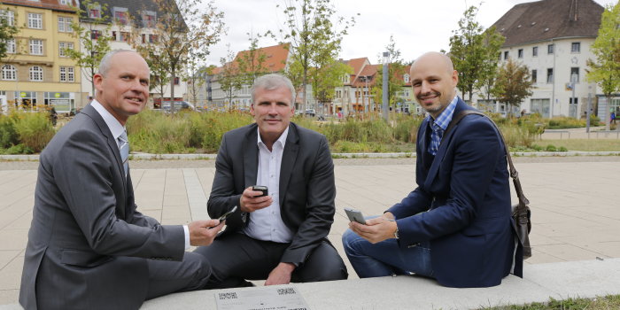 Drei Männer mit Smart-Phones in der Hand sitzen neben einer GPS-Referenzplatte, im Hintergrund sieht man die Parkanlage Hirschgarten. 