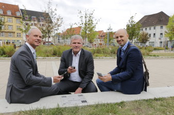 Drei Männer mit Smartphones in der Hand sitzen neben einer GPS-Referenzplatte, im Hintergrund sieht man die Parkanlage Hirschgarten. 
